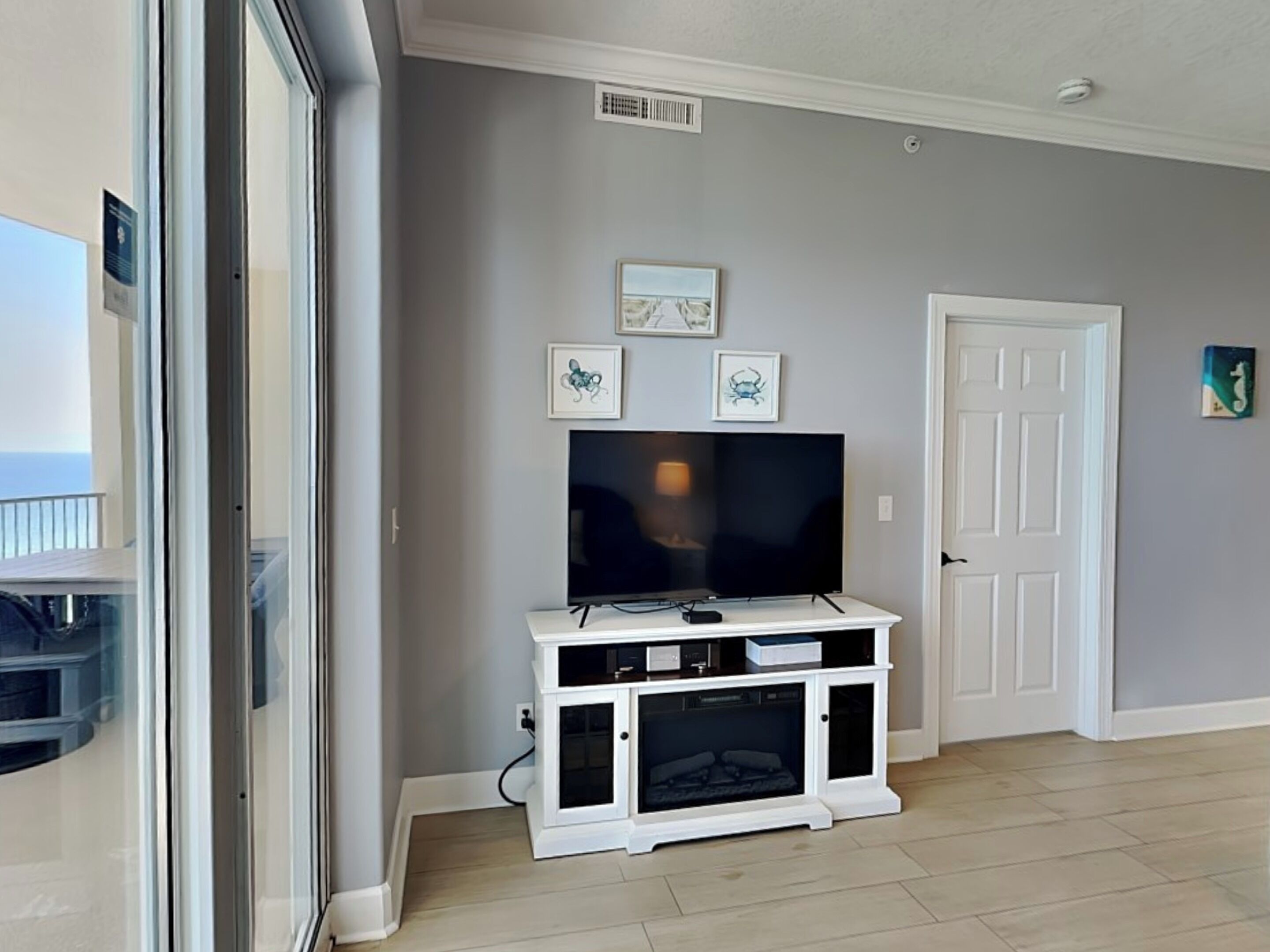 A living room with a flat-screen TV on a white entertainment unit, a door, and a window with a view.
