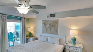 Una habitación de hotel con una cama, dos mesitas de noche, un mapa del mundo en la pared, un ventilador de techo y un balcón con una silla.