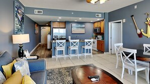 Un salon moderne avec un canapé, une table basse et un téléviseur. Il y a également un coin cuisine avec un bar et des chaises blanches.