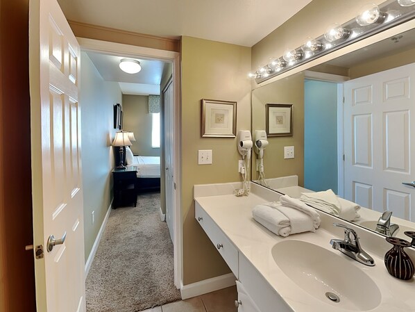 Un baño moderno con un mueble de tocador espacioso, un lavabo y un espejo con iluminación. Se ve una puerta que conduce a un dormitorio y a un pasillo.