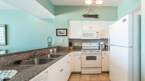 A compact kitchen with white cabinets, a double sink, a microwave, a stove, and a refrigerator. - Shores of Panama #1111 | Pool + Gym! (Panama City Beach)
