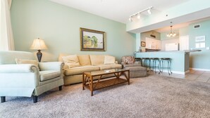 A living room with a sofa, armchairs, a coffee table, and a kitchen area with a bar and stools. - Shores of Panama #1111 | Pool + Gym! (Panama City Beach)