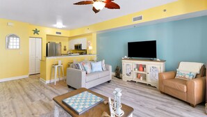 Sala de estar con sofá, mesa de centro y televisor. Zona de cocina con refrigerador, microondas y mesa de comedor. La habitación tiene ventilador de techo y ventana.