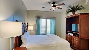 A neatly arranged bedroom with a bed, bedside lamps, a ceiling fan, a television on a wooden cabinet, and a window with curtains. - Shores of Panama #1510 | Lagoon Pool! (Panama City Beach)