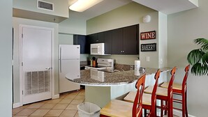 A kitchen with a central island, a microwave, a refrigerator, and a trash can. There are chairs around the island and a sign on the wall that says "WINE BAKERY". - Shores of Panama #1510 | Lagoon Pool! (Panama City Beach)