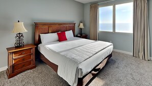 A bedroom with a bed, two bedside tables, a ceiling fan, a window with curtains, and a view of the ocean.