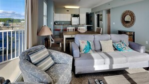 A living room with a grey sofa, patterned armchair, and a view of the kitchen and balcony.
