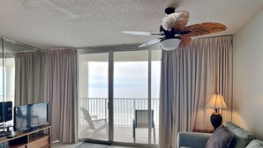 Ein modernes Hotelzimmer mit einer großen Glasschiebetür, die auf einen Balkon führt, einem Flachbildfernseher, einem Sofa, einem Couchtisch und einem Deckenventilator mit Blattformdesign.