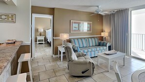 A modern living room with a sofa, armchair, and a dining area. There is a ceiling fan, a window with curtains, and a door leading to another room. - Long Beach III #905 | Incredible Gulf Views (Panama City Beach)