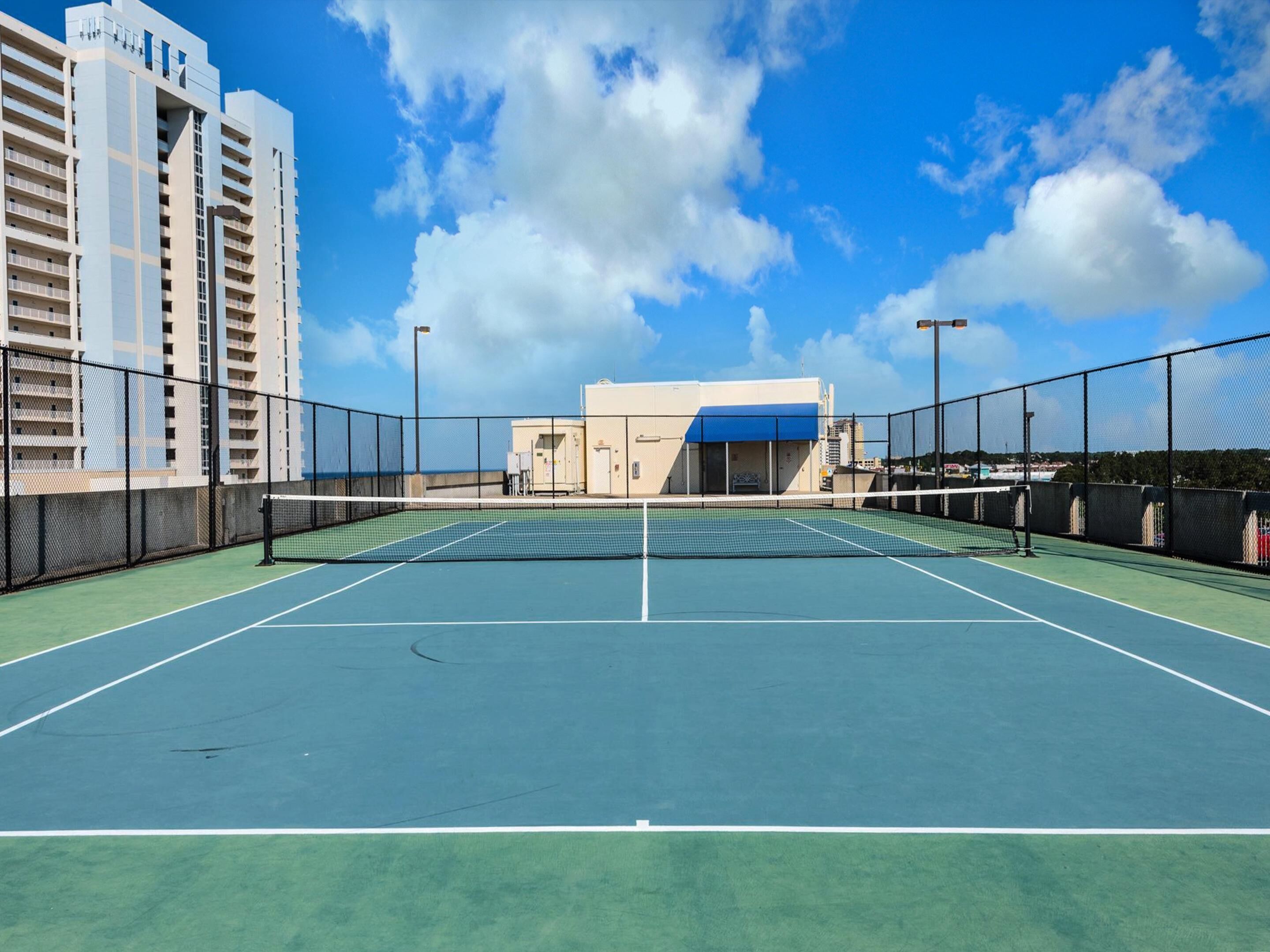 Een buitenterrein voor tennis met een net, omgeven door een hek, naast een gebouw met meerdere verdiepingen.