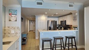 A modern kitchen with a central island, bar stools, and a tiled backsplash. - Majestic Beach Towers II #1902 | Luxe Gulf Front + Amenities (Panama City Beach)