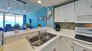 A kitchen with white cabinets, a stainless steel sink, a coffee maker, and a clock on the wall. - Majestic Beach Towers II #707 | Gulf Front + Resort Amenities (Panama City Beach)