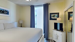 A hotel room with a bed, a TV, a nightstand, and a mirror. - Majestic Beach Towers I #1015 | Gulf Front + Resort Amenities (Panama City Beach)