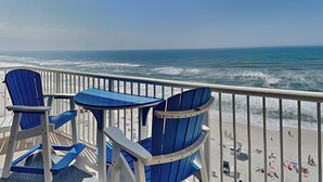 A balcony with blue Adirondack chairs and a small table, overlooking a beach with people and umbrellas. - Majestic Beach Towers I #1015 | Gulf Front + Resort Amenities (Panama City Beach)