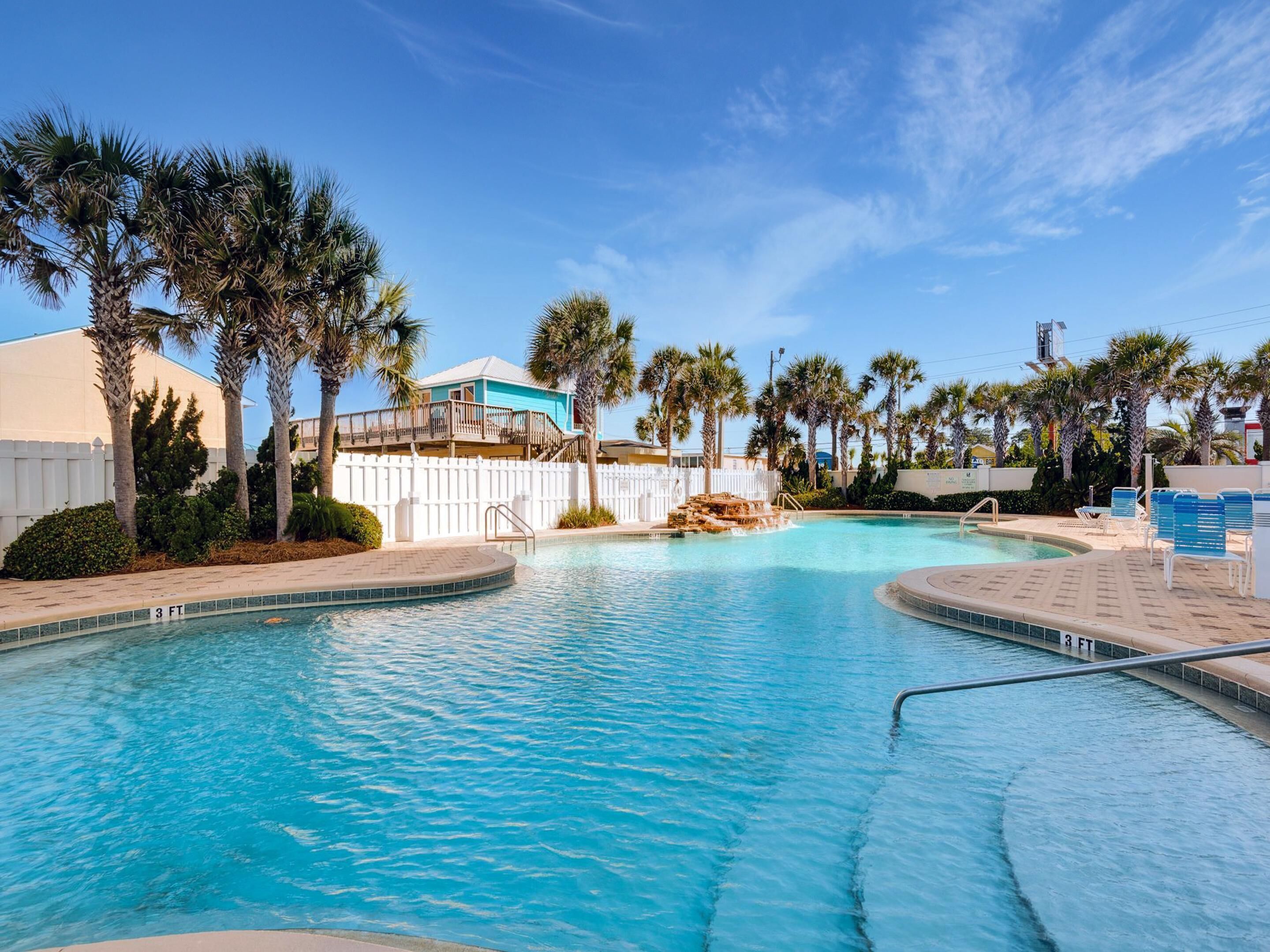 A swimming pool with a white fence, palm trees, and a building with a blue roof.