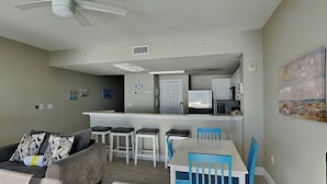 A modern living room with a grey sofa, blue chairs, a white table, and a kitchen area with a fridge and microwave. - Majestic Beach Towers I #1410 | Gulf Front + Amenities (Panama City Beach)