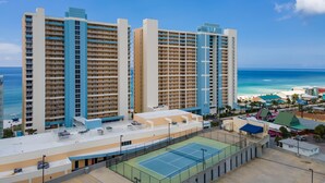 A high-rise hotel with multiple balconies, a tennis court, and a view of the beach.