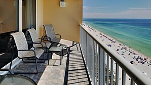A balcony with three chairs and a table, overlooking a beach with umbrellas and people.