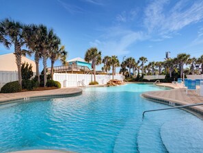 A swimming pool with a white fence, palm trees, and a building with a blue roof. - Majestic Beach Towers I #1608 | Gulf Front + Amenities (Panama City Beach)