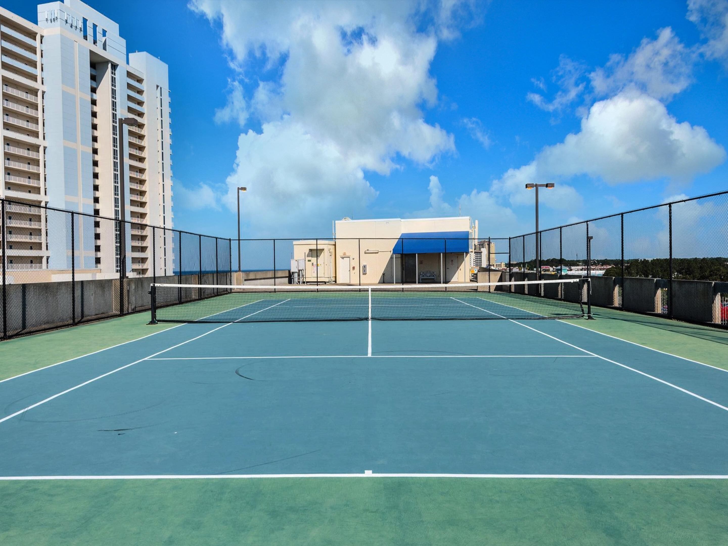 An outdoor tennis court with a net, surrounded by a fence, and adjacent to a building with multiple floors.