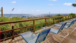 A wooden deck with blue chairs, a railing, and a view of the beach with umbrellas and the ocean.