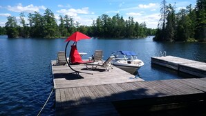 A wooden dock with lounge chairs and a red umbrella, a boat docked at the pier, and a forested shoreline in the background.