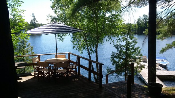 A wooden deck with a table and chairs, a striped umbrella, and a view of a lake with a dock and boat.