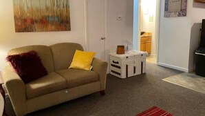 A living room with a beige sofa, a red and yellow throw pillow, a white side table with a basket, and a painting of trees on the wall. - Vacation Condo #5 2BR with full kitchen, walk to river and shops, pets welcome (Hardy)