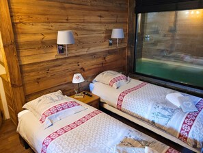 A room with wooden walls, two beds with patterned bedding, bedside lamps, a nightstand with a book and remote, and a window with a view. - Chalet de prestige 18 people swimming pool jacuzzi sauna (Vars)
