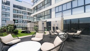 An outdoor seating area with wicker chairs and a round table, surrounded by a modern building with large windows and a covered entrance.
