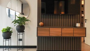 A modern living area with a large flat-screen TV mounted on a wooden panel wall, two potted plants on metal stands, and a round mirror on the wall.