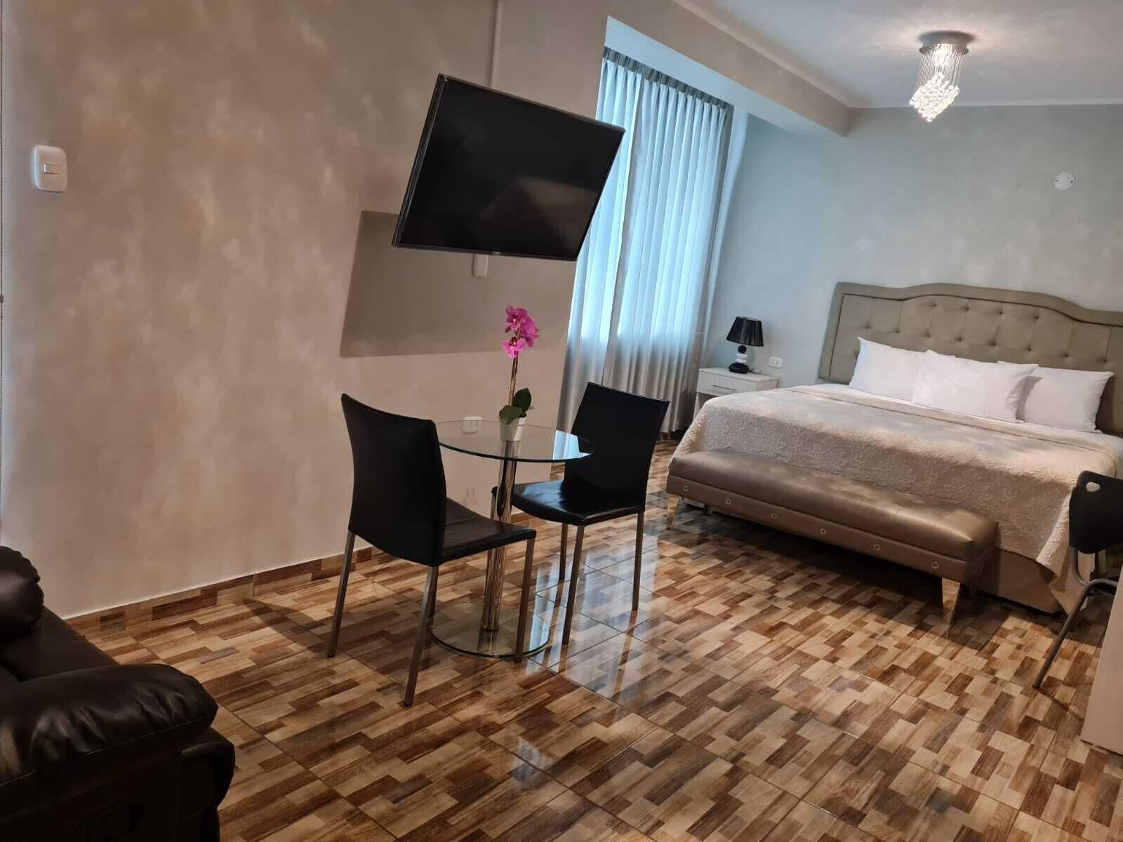 A hotel room with a bed, a TV mounted on the wall, a desk with two chairs, a small table with a flower, and a chandelier.