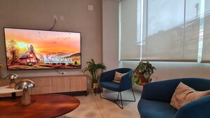 Ein modernes Wohnzimmer mit an der Wand montiertem Fernseher, einem Holzschrank, einem runden Couchtisch, zwei blauen Sesseln und einem großen Fenster mit Jalousien.