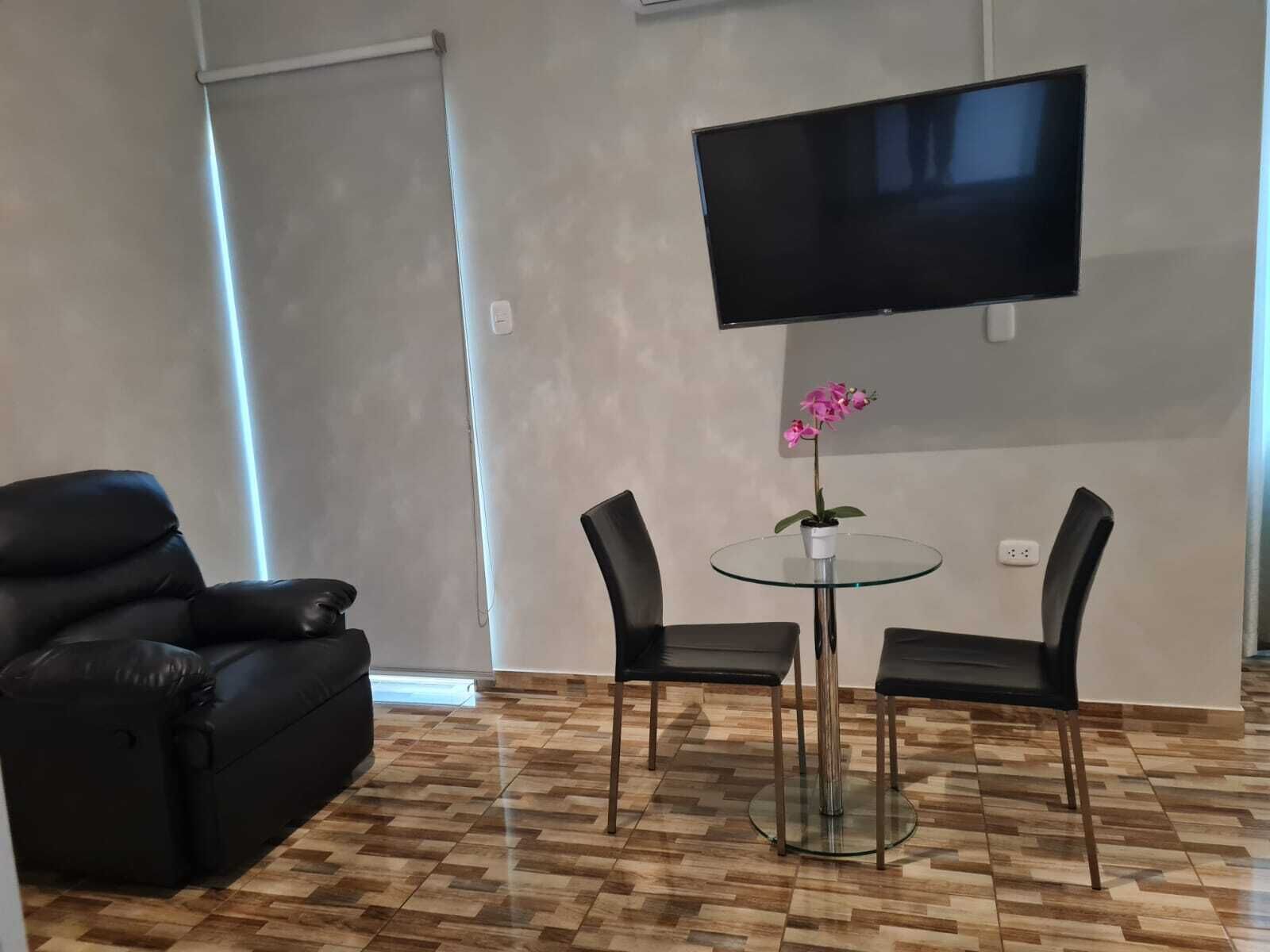 A modern living room with a black leather sofa, a glass table with two chairs, a potted orchid, and a mounted flat-screen TV.