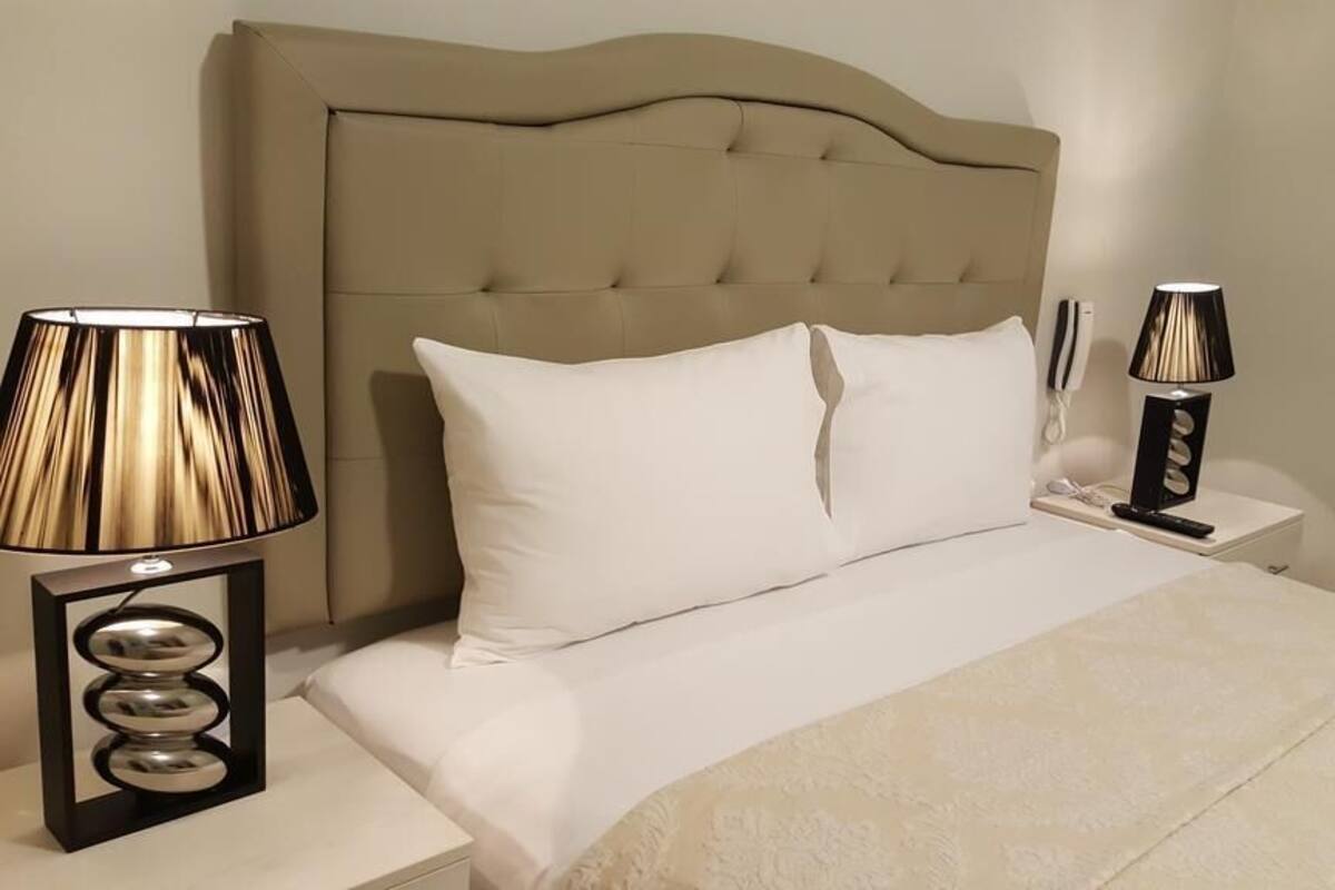 Un lit bien fait, avec du linge de lit blanc, une tête de lit recouverte d’un tissu beige et deux tables de chevet avec des lampes.