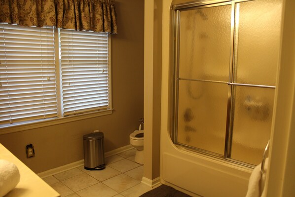 A bathroom with a shower, toilet, and window with blinds.
