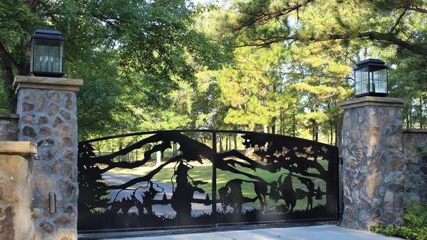 A gated entrance with a metal gate featuring a tree and animal silhouette design, flanked by stone pillars with lanterns. - Farmhouse on Beautiful Bison Farm (Opelika)