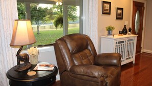 A leather armchair by a window with a lamp, a small table with decorative items, and a white console table with framed pictures and a vase. - Farmhouse on Beautiful Bison Farm (Opelika)