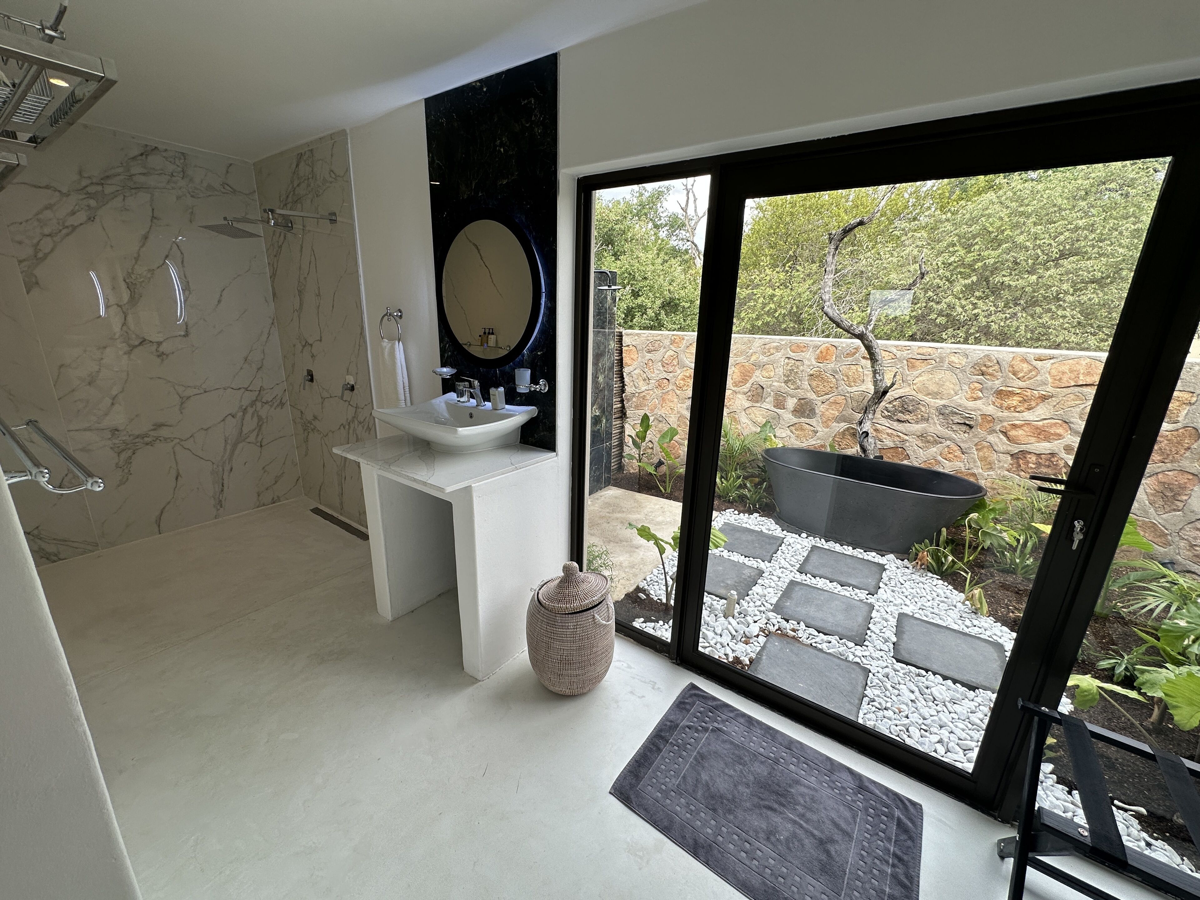 Een moderne badkamer met een marmeren douche, een wastafel met een grote spiegel en uitzicht op een tuin door een glazen deur.