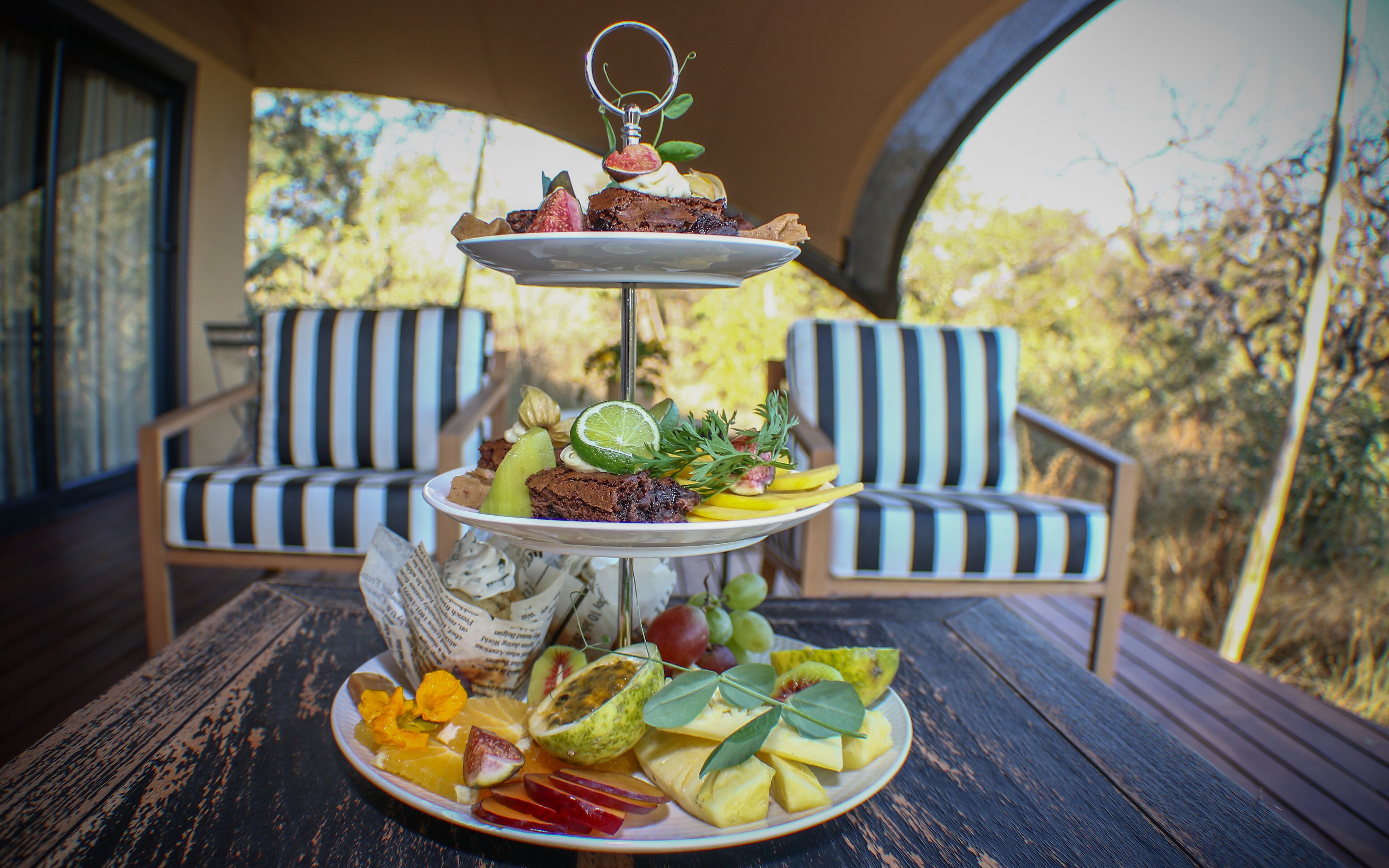 Un soporte escalonado para postres con diversas frutas y un pastel sobre una mesa de madera, rodeado de sillas a rayas.