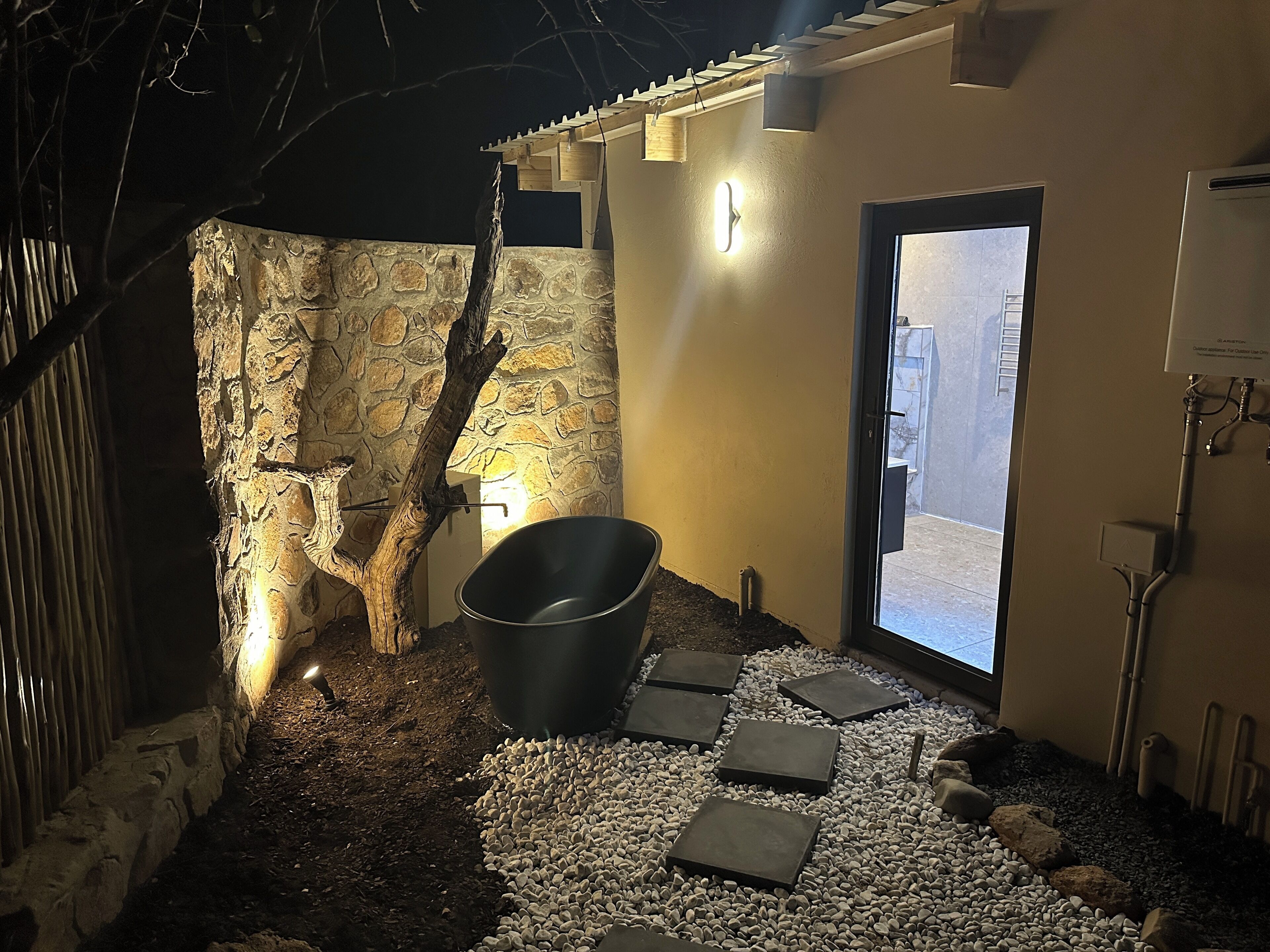 A stone wall with a rustic tree trunk, a black bucket, and a pathway leading to a door.