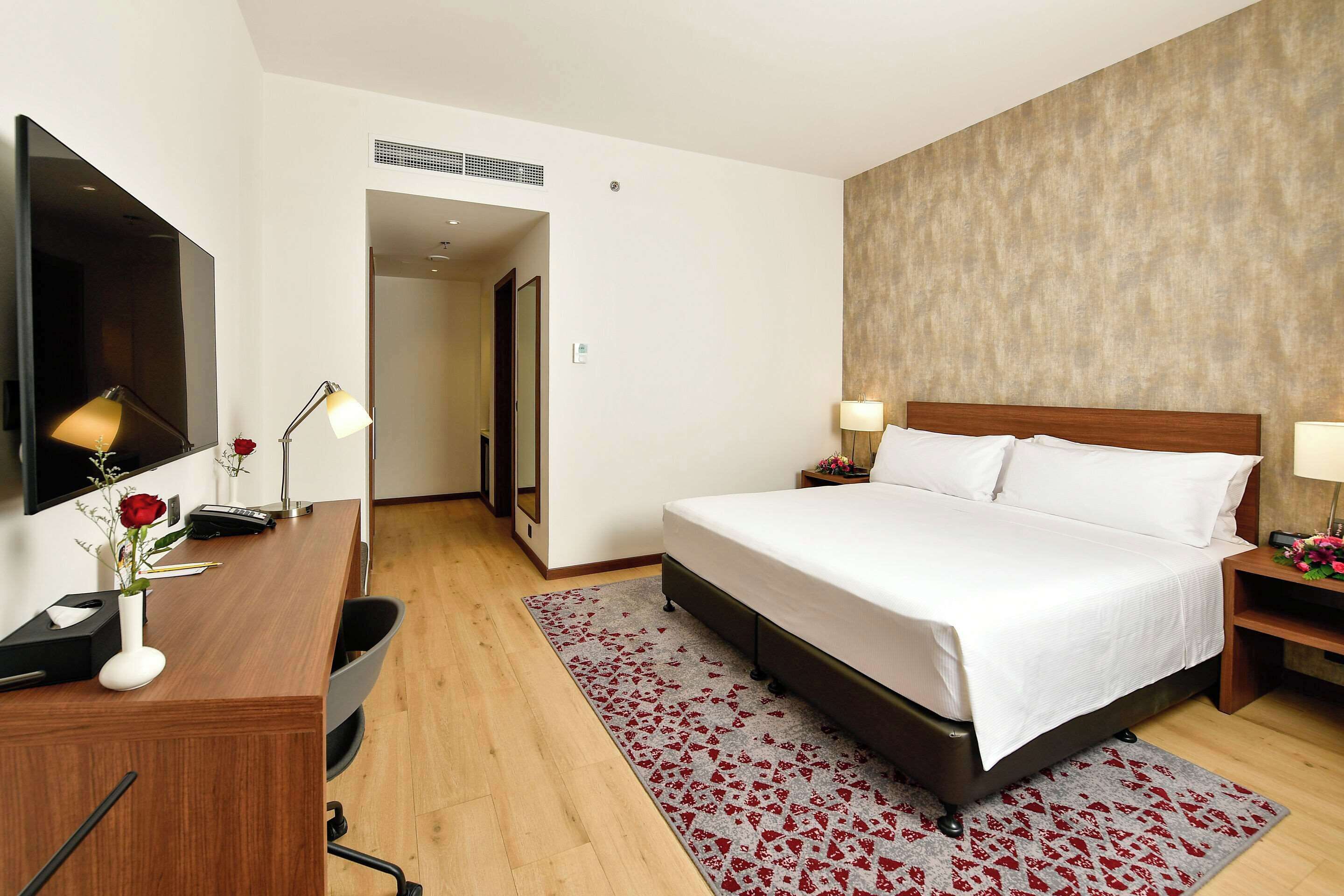 Een hotelkamer met een groot bed, een bureau met een flatscreen televisie, twee bedlampen en een vloerkleed met een rood-wit patroon.
