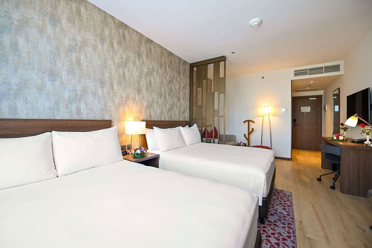 A hotel room with two beds, a desk, a lamp, and a mirror.