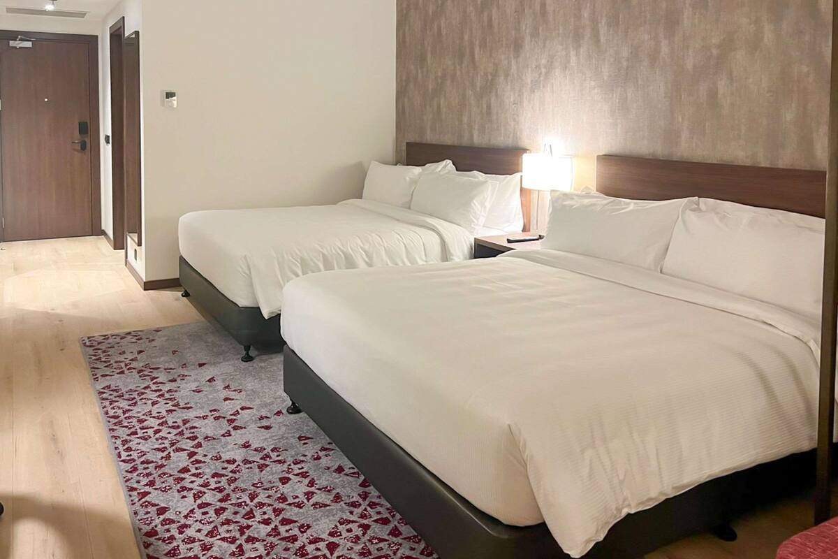 Une chambre d’hôtel avec deux lits, une tête de lit en bois, une table de chevet et un tapis à motifs rouges et blancs.