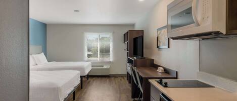 A compact hotel room with two single beds, a kitchenette with a microwave, stove, and dishwasher, a TV, and a window with blinds.
