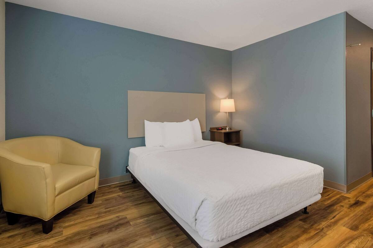 Una habitación de hotel con una cama, una silla y una mesita de noche con lámpara.