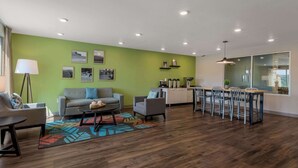 Ein modernes Wohnzimmer mit einer grünen Akzentwand, einem grauen Sofa, einem Essbereich mit Tisch und Stühlen sowie einer Küche im Hintergrund.