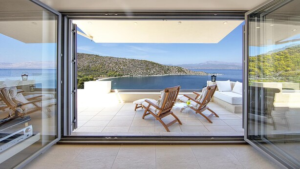 A modern balcony with a glass door, wooden chairs, a white sofa, and a view of a mountainous landscape and sea. - Crystal View Villa by Unique Villas Collection (Corinth)