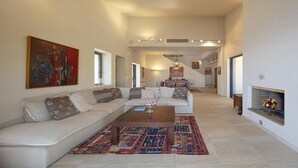 A spacious living room with a white sofa, a coffee table, and a colorful rug. There is a fireplace with a flower arrangement on the right and a painting on the left. - Crystal View Villa by Unique Villas Collection (Corinth)