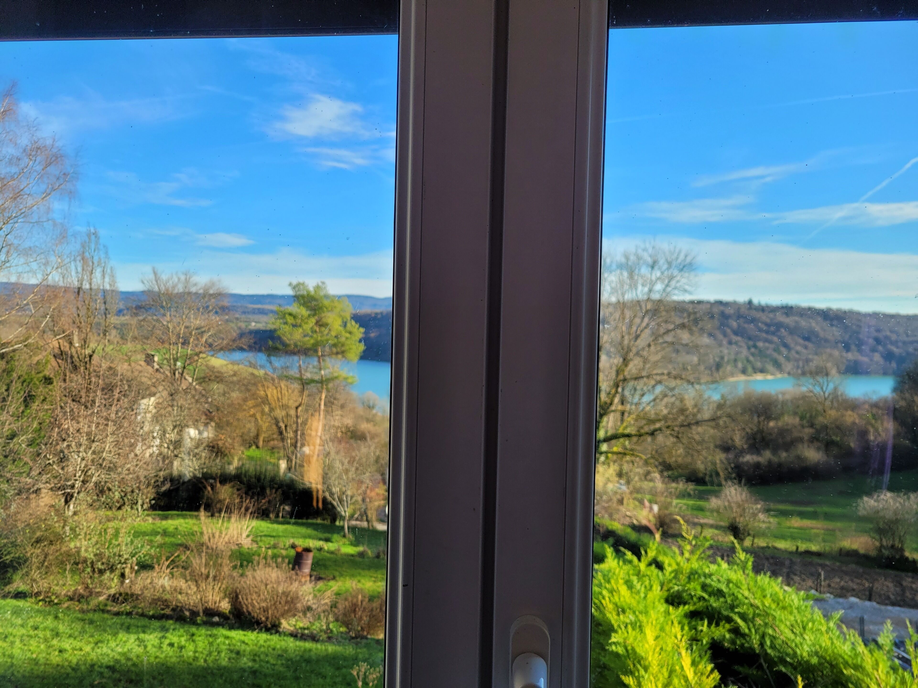 A view through a window with a clear sky, a lake, and surrounding greenery.